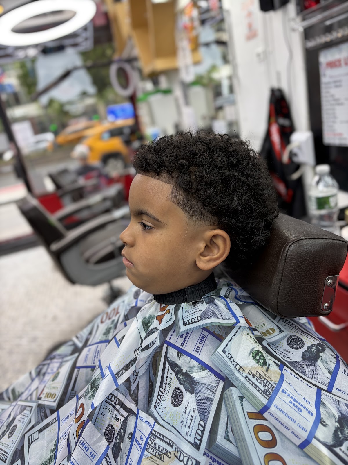Young boy seated in a barber chair with curly top and low fade, dollar bill print cape